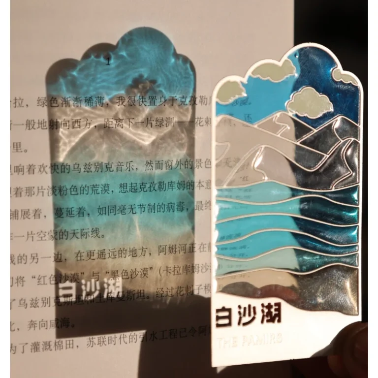 Metal bookmark with turquoise wave and landscape enamel details placed on an open book.