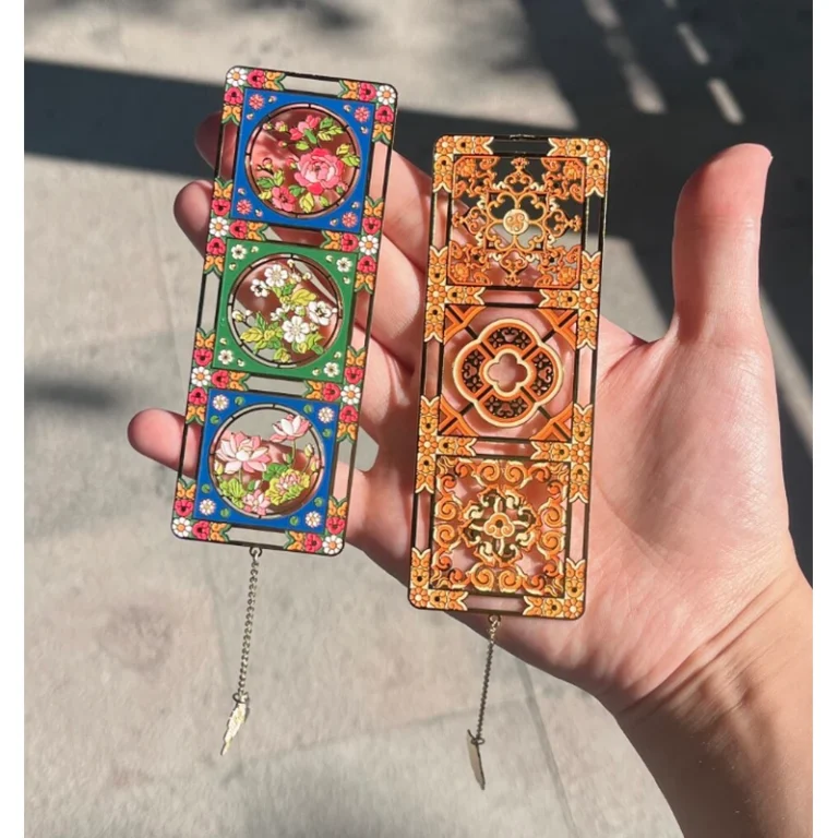 Two colorful rectangular metal bookmarks with floral window and ornamental clock designs, held in a hand.