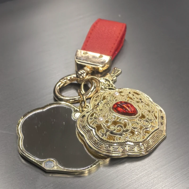 Gold-tone sculpted folding keychain mirror with red decorative detail and red strap.