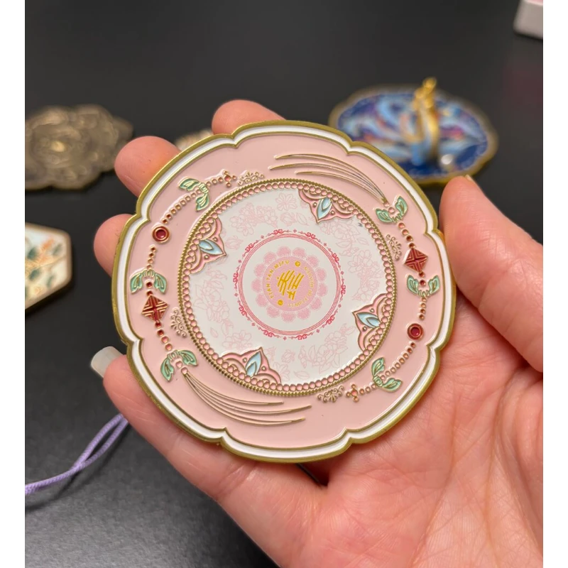 Pink round enamel compact mirror with decorative motifs and red accents, held in a hand.