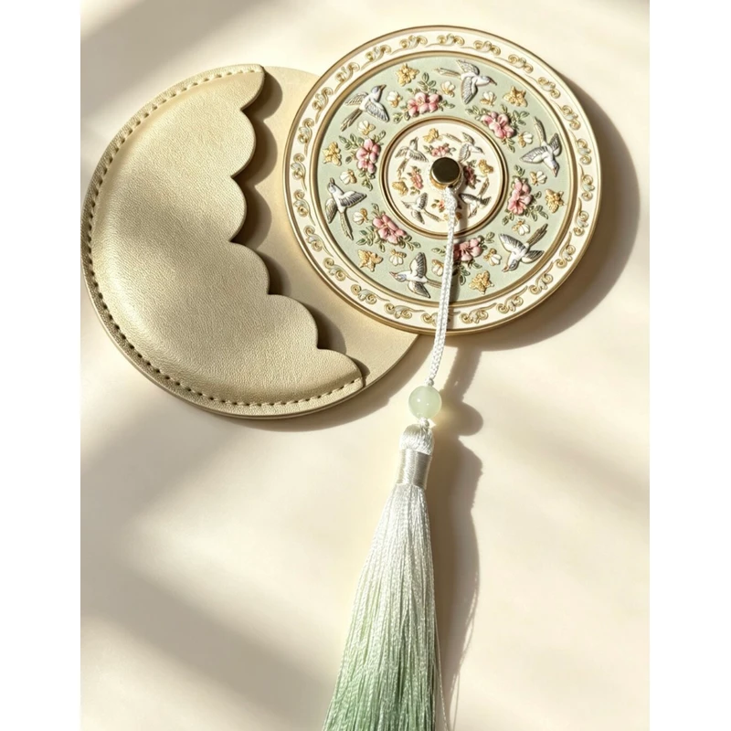 Gold-tone and white floral handheld mirror with tassel beside round presentation packaging.