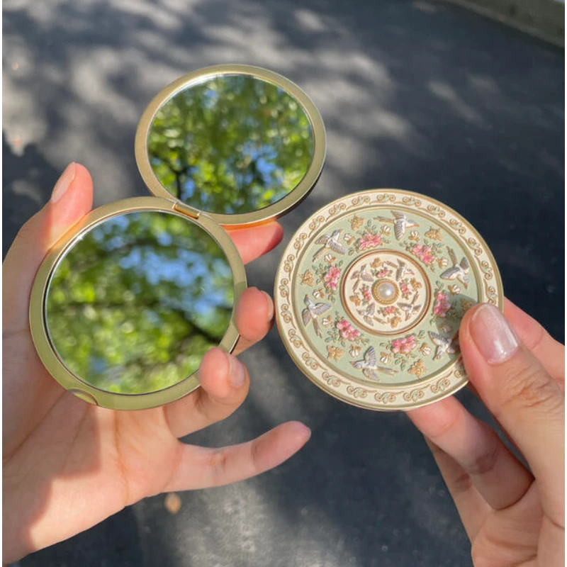 Open folding mirror with a round floral decorated cover, held in two hands.